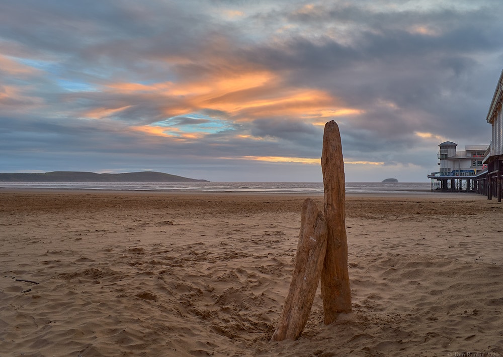 Ein Bild mit hoher Qualität, das das Endprodukt des im vorherigen Screenshot gezeigten Bildes von zwei stehenden Treibholzblöcken an einem Strand bei Sonnenuntergang zeigt, mit hohem Farbkontrast und Objektschärfe.