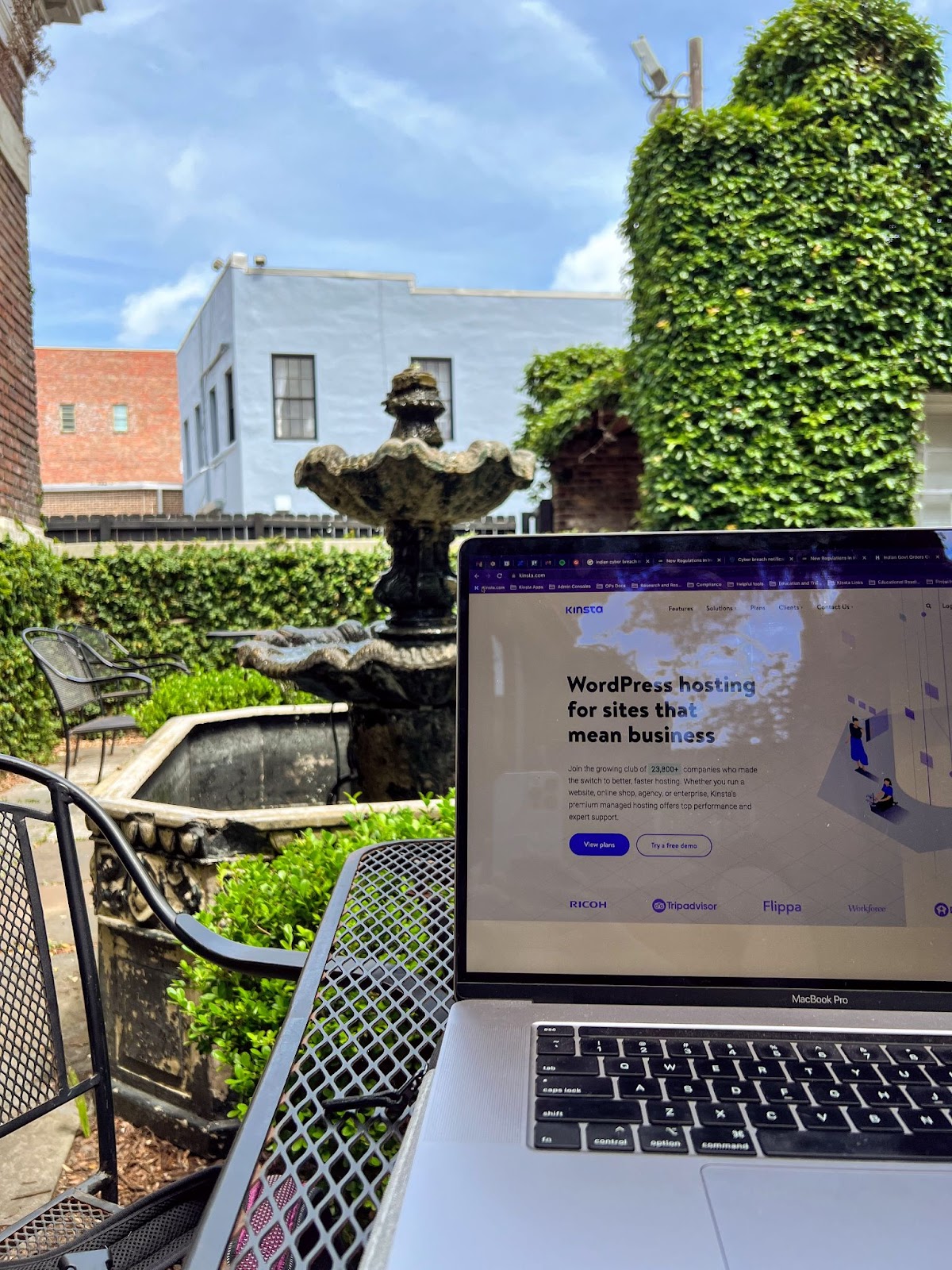 A laptop outdoors on a coffee table with a view of a fountain and ivy-covered walls.