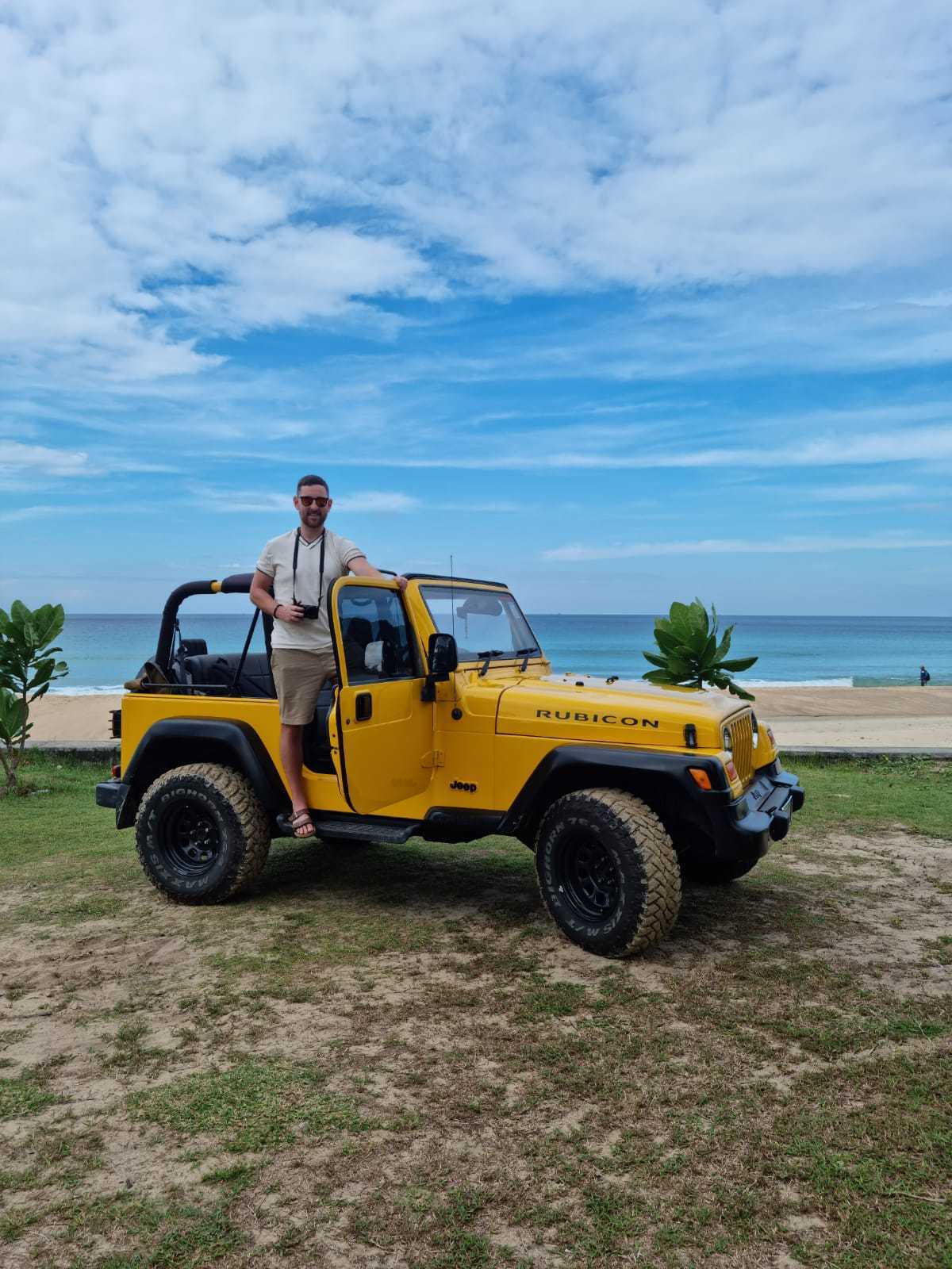 A man standing at the open door of a yellow Jeep Rubicon with a view of the ocean in the background.