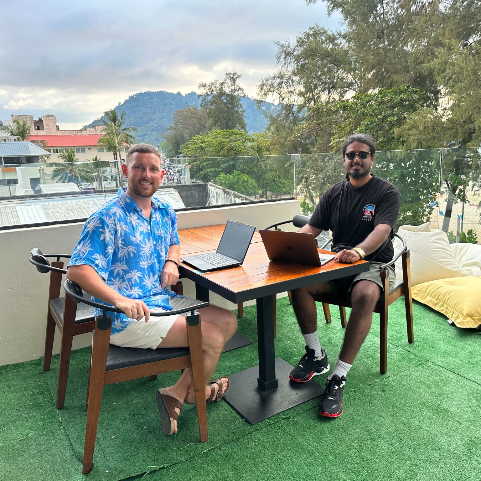 Two smiling men sitting with laptops at a table in an outdoor rooftop area.