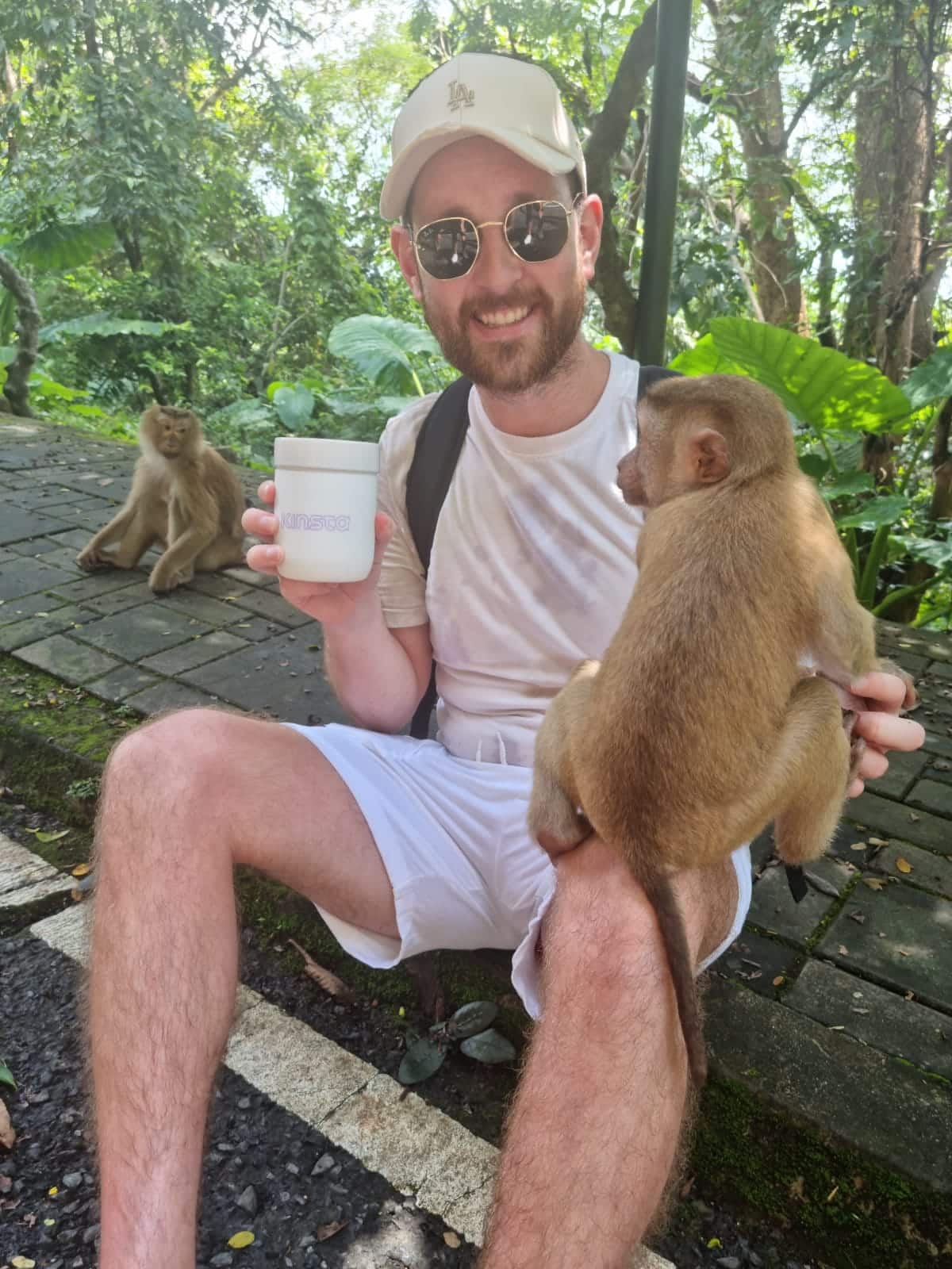 A smiling man in a white baseball cap sitting in a tropical environment, holding a beverage cup in his right hand and touching a brown monkey that's balancing on his left knee with his left hand.