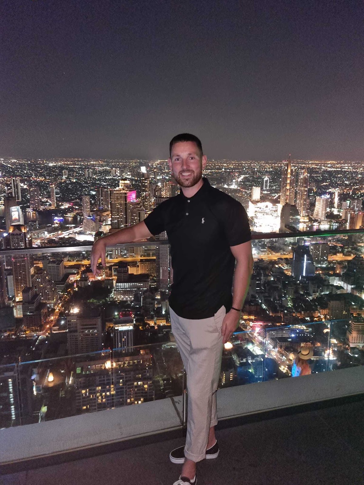 A smiling man standing on a high balcony above a lit city, with his right arm resting on the balcony's glass railing.